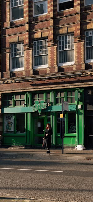 Exterior view of a traditional brick building with large windows on upper floors, casting shadows on the facade. The lower storefront features bright green painted wooden framing and large glass windows revealing a commercial space inside, with a woman dressed in black walking past the entrance on the sidewalk. The scene is well-lit by natural daylight, and the street has minimal traffic. The storefront signage indicates a retail or service establishment, aligned with cleaning or maintenance services as promoted by Maida Vale Carpet Cleaners in their page about avoiding hidden charges in cleaning quotes.