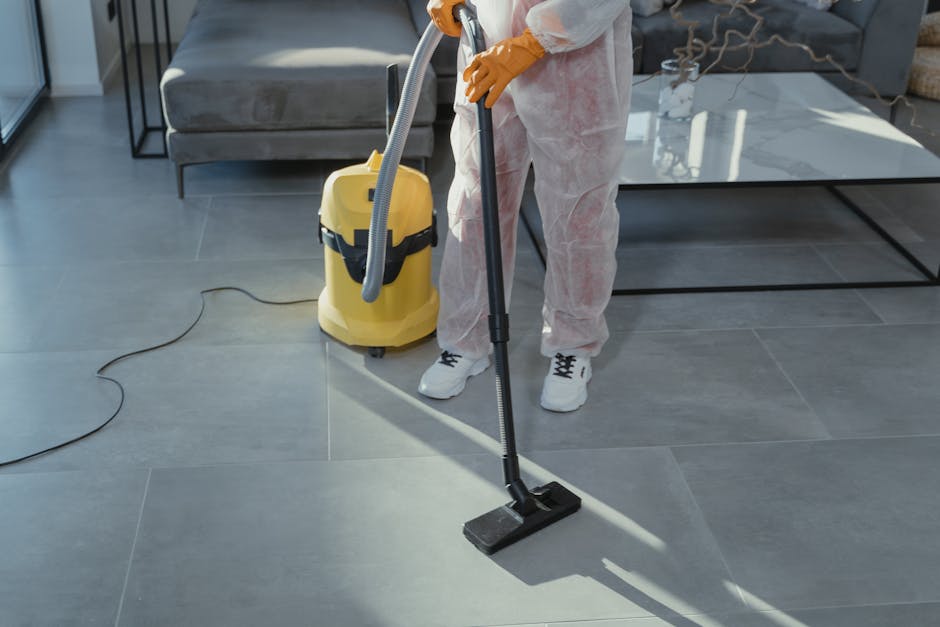 A professional cleaner wearing a white protective suit, mask, and orange gloves operates a vacuum cleaner on a light-colored tiled floor in a modern, well-lit room with large windows. The room has a m