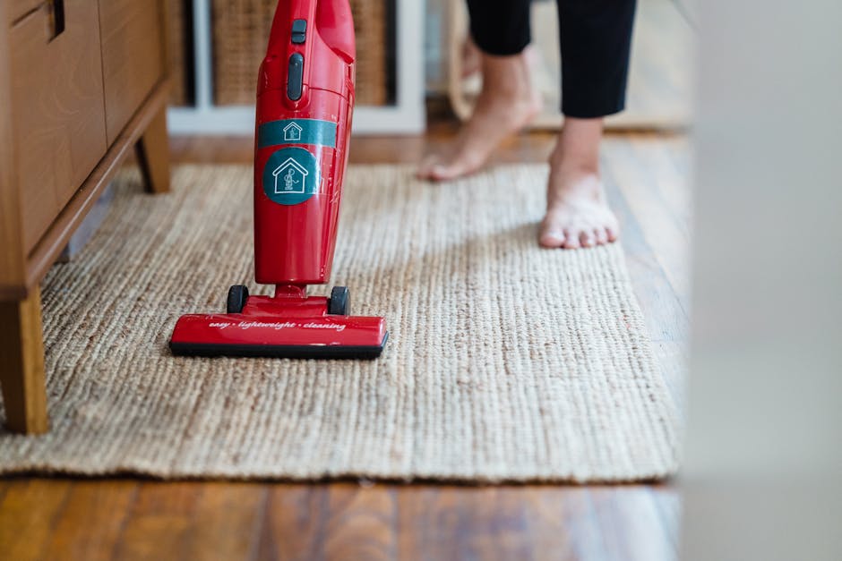 A person using a red handheld vacuum cleaner to perform surface cleaning on a beige woven rug in a residential living room. The vacuum features a blue power button and a transparent dust container, and is positioned on the edge of the rug, which covers a wooden floor. The person's bare feet are visible, and they are wearing black pants. The room is well-lit, with natural light coming from the side, emphasizing the clean and tidy condition of the rug, as part of Maida Vale Carpet Cleaners' domestic cleaning and sanitisation services for Little Venice homes. The scene highlights the importance of regular deep cleaning to maintain hygiene and preserve the appearance of home surfaces.