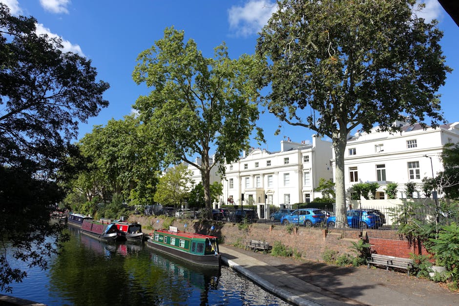 A bright, sunny day along a residential canal in Little Venice, Maida Vale. The image features a clear blue sky with a few white clouds, and the canal's calm water reflecting the surroundings. On the left, partially visible, are lush green trees with dense foliage extending over the water. Along the canal edge, there are several narrowboats painted in various colors, including maroon and green, moored securely. On the right side, a row of elegant white terraced houses with large windows and decorative architectural details face the canal. A few cars are parked along the street, and there are small gardens with well-maintained plants and shrubs. The scene is well-lit, highlighting the cleanliness of the environment and the vibrant greenery, reflecting a typical picturesque residential area in Maida Vale. Maida Vale Carpet Cleaners recommends surface cleaning and maintenance of such areas to preserve their appeal and hygiene.