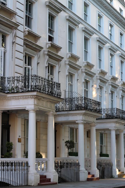 A wide street lined with elegant white residential buildings featuring detailed architectural facades, large windows, and balconies. The street is shaded by a leafless tree on the left, with parked ca