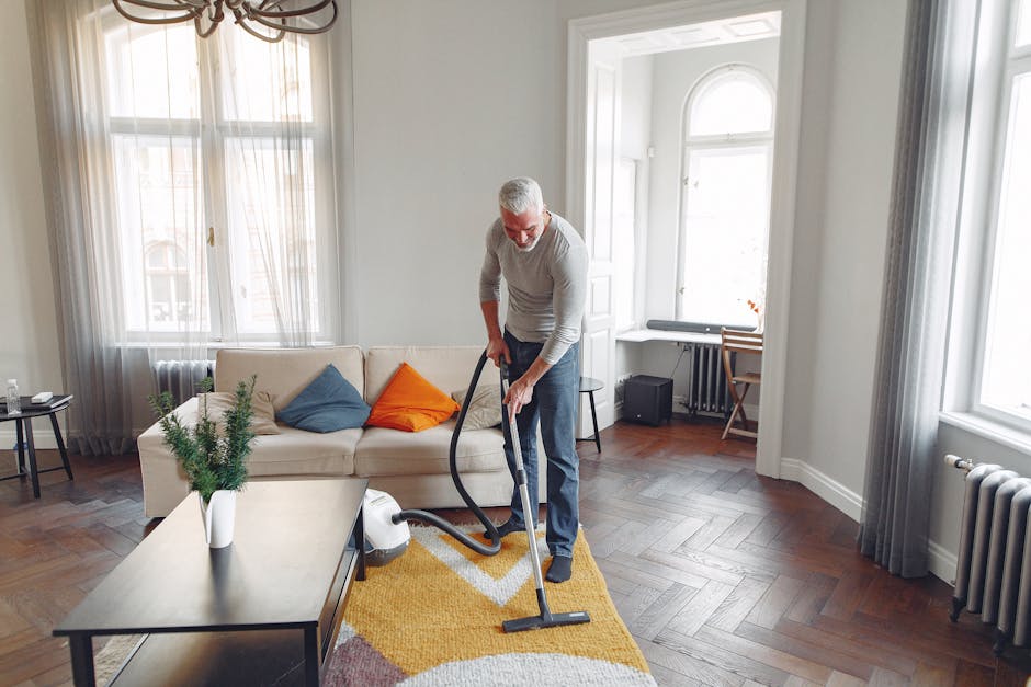 A person kneeling on a patterned carpet in a room with warm lighting, opening a yellow and black portable vacuum cleaner with a flexible hose attachment. The individual is wearing a beige jacket and b