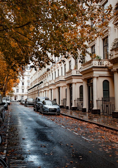 A residential street in Maida Vale with a row of elegant white terraced buildings featuring ornate architectural details and balconies. The pavement appears wet, reflecting the overcast sky, with scat